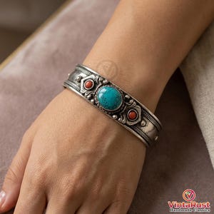 May include: A silver cuff bracelet featuring a central oval turquoise stone flanked by two smaller round coral-colored stones. The metal surface displays intricate embossed patterns, decorative metal beads, and etched line details throughout.