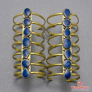 May include: Two brass arm cuffs, each featuring a vertical row of blue gemstone accents. The cuffs have multiple curved bands and a central decorative element. The gemstones are oval and teardrop-shaped. The background is a neutral gray.