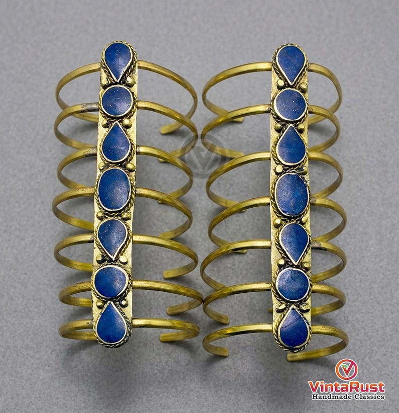 May include: Two brass arm cuffs, each featuring a vertical row of blue gemstone accents. The cuffs have multiple curved bands and a central decorative element. The gemstones are oval and teardrop-shaped. The background is a neutral gray.