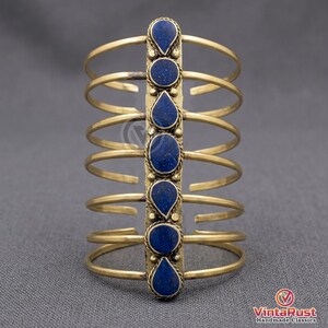 May include: A gold-toned arm cuff featuring a vertical row of six dark blue gemstones. The cuff has multiple parallel bands connected by a central decorative element. The gemstones are oval and teardrop-shaped, set in a detailed metalwork design.