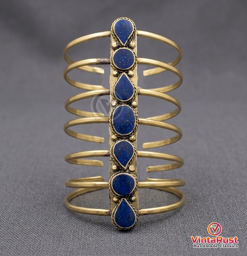 May include: A gold-toned arm cuff featuring a vertical row of six dark blue gemstones. The cuff has multiple parallel bands connected by a central decorative element. The gemstones are oval and teardrop-shaped, set in a detailed metalwork design.