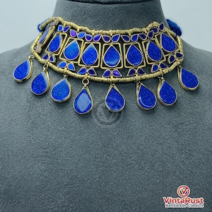 May include: A gold-toned choker necklace with vibrant blue gemstone accents. The necklace features a series of square and teardrop-shaped blue stones, creating a striking contrast against the gold metal. The necklace is displayed on a gray mannequin head.