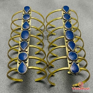 May include: Two sets of brass arm cuffs, each featuring a central vertical band adorned with six dark blue, teardrop-shaped stones. The cuffs have multiple curved brass bands extending outwards. The image is set against a gray background.