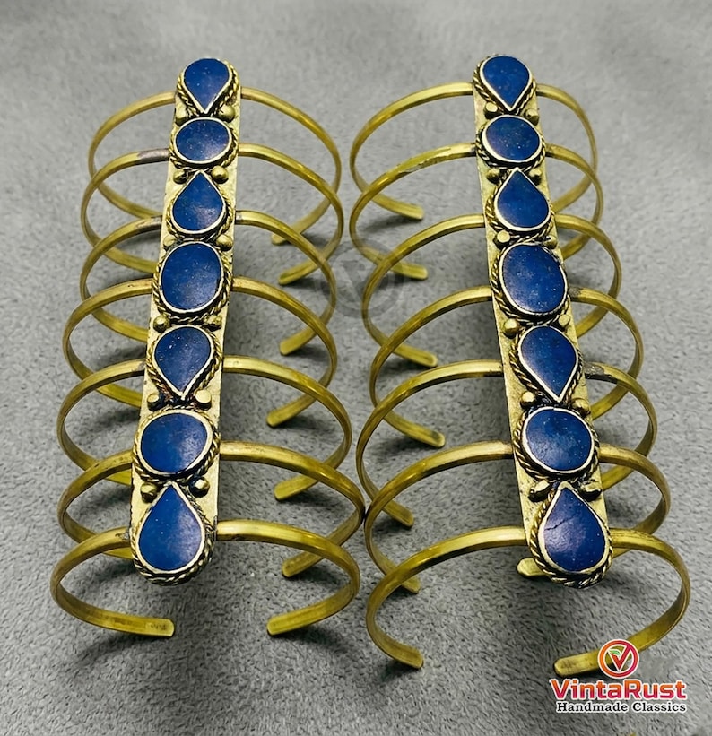 May include: Two sets of brass arm cuffs, each featuring a central vertical band adorned with six dark blue, teardrop-shaped stones. The cuffs have multiple curved brass bands extending outwards. The image is set against a gray background.