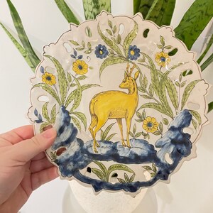May include: A white ceramic plate featuring a yellow deer standing on a blue and green rock. The plate is decorated with yellow and blue flowers and green leaves. The plate has a scalloped edge and is hand-painted.