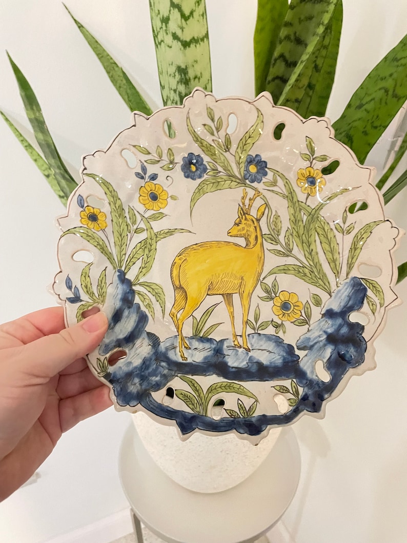 May include: A white ceramic plate featuring a yellow deer standing on a blue and green rock. The plate is decorated with yellow and blue flowers and green leaves. The plate has a scalloped edge and is hand-painted.