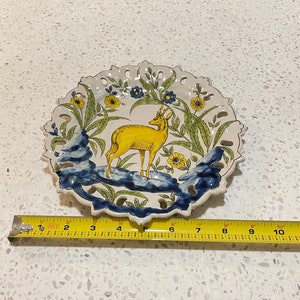 May include: A white ceramic plate featuring a yellow deer within a blue and green floral design. The plate has a scalloped edge and measures approximately 18 cm in diameter.
