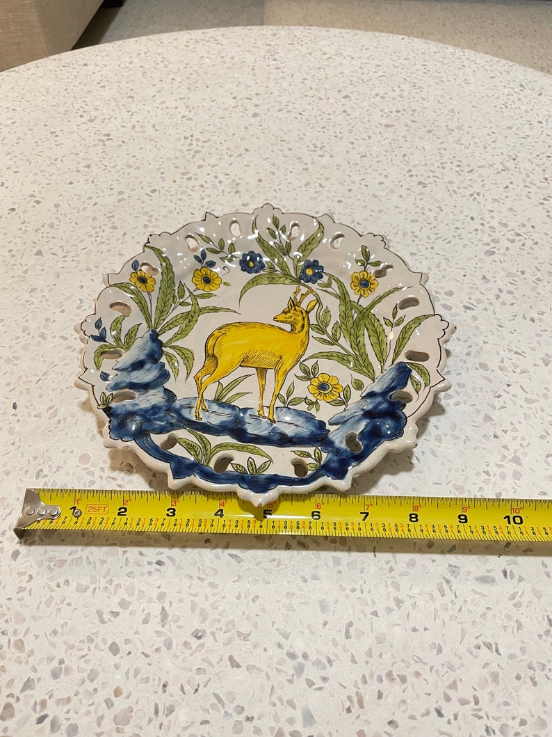 May include: A white ceramic plate featuring a yellow deer within a blue and green floral design. The plate has a scalloped edge and measures approximately 18 cm in diameter.