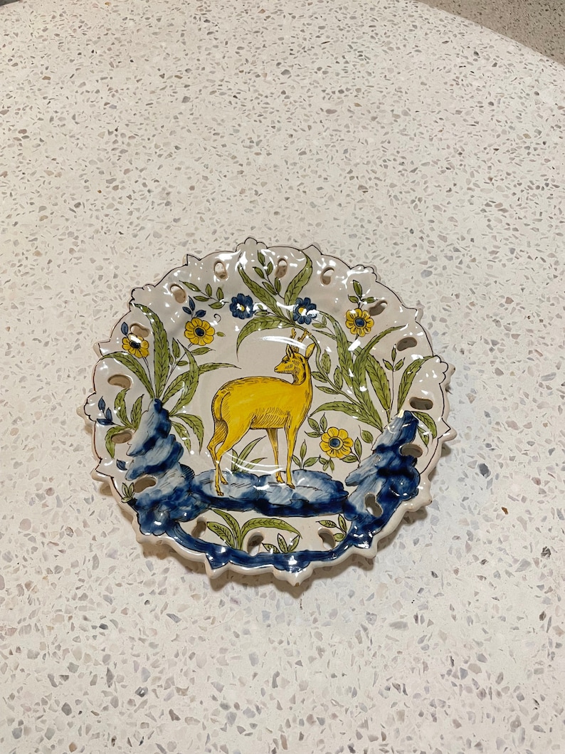 May include: A white ceramic plate featuring a yellow deer standing on a blue and green rock. The plate has a scalloped edge and a floral design.