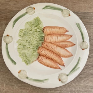 May include: White ceramic plate with a hand-painted design of carrots and green onions. The carrots are arranged in a fan shape and the onions are around the edge of the plate.