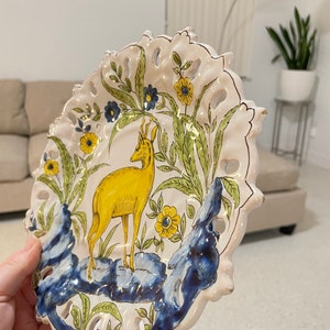 May include: A white ceramic plate featuring a yellow deer, green foliage, and blue and yellow flowers. The plate has a scalloped edge and is decorated with a gold trim.