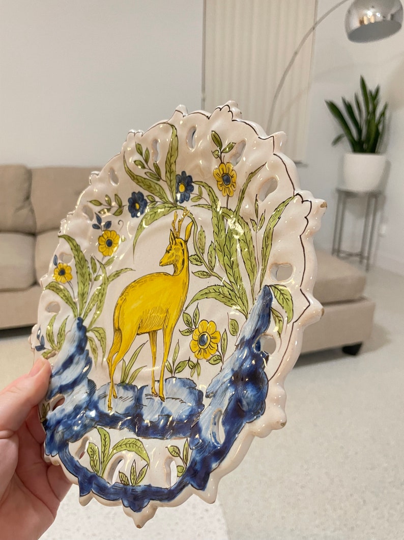 May include: A white ceramic plate featuring a yellow deer, green foliage, and blue and yellow flowers. The plate has a scalloped edge and is decorated with a gold trim.