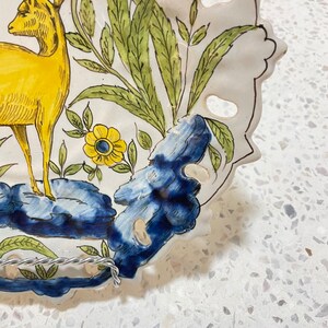 May include: A white ceramic plate with a blue and yellow floral design. The plate features a yellow deer with antlers and a blue and white floral design. The plate has a scalloped edge and is decorated with holes.