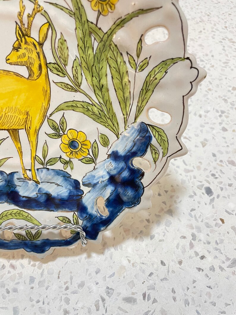 May include: A white ceramic plate with a blue and yellow floral design. The plate features a yellow deer with antlers and a blue and white floral design. The plate has a scalloped edge and is decorated with holes.