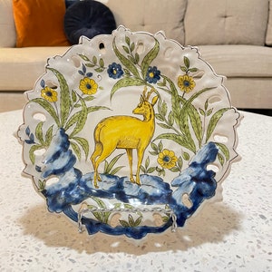 May include: A white ceramic plate with a blue and yellow floral design and a yellow deer in the centre. The plate has a scalloped edge and is decorated with a delicate lace pattern.