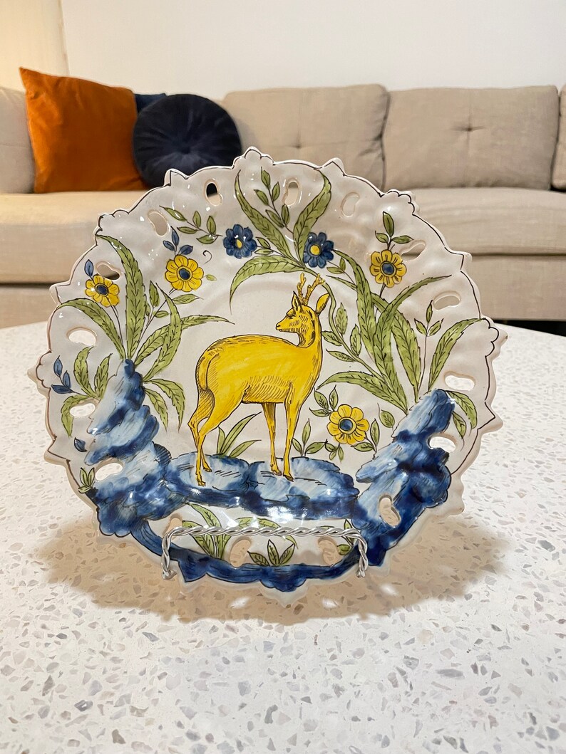 May include: A white ceramic plate with a blue and yellow floral design and a yellow deer in the centre. The plate has a scalloped edge and is decorated with a delicate lace pattern.
