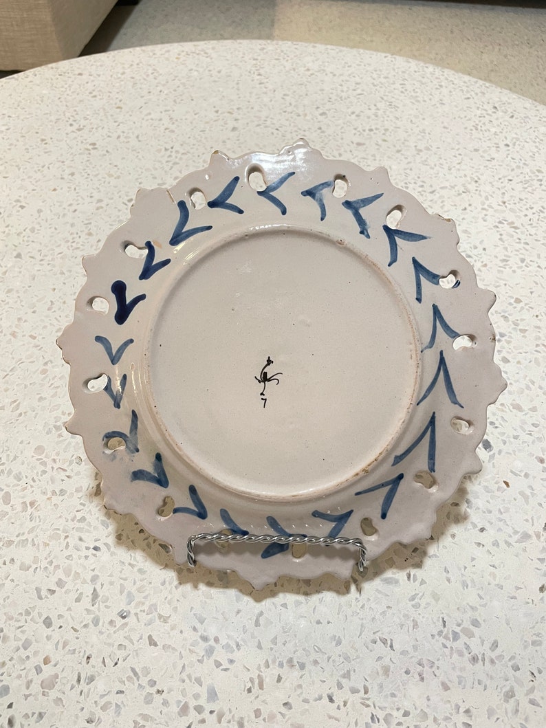 May include: White ceramic plate with a blue and white design of arrows around the edge. The plate has a scalloped edge and is decorated with a blue and white floral design. The back of the plate has a small black mark.