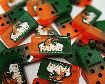 May include: A collection of rectangular dominoes featuring a translucent orange and green resin finish. Each piece displays the FAMU Rattlers logo, which includes a snake illustration and the text FAMU RATTLERS, alongside standard black pips.