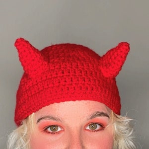 May include: Red crocheted hat with devil horns. The hat is made of a soft, textured yarn and has a comfortable fit.