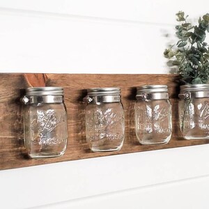 May include: A rustic wooden wall shelf with four clear glass mason jars attached with metal clips. The jars are empty and ready to be used for storage or decoration.