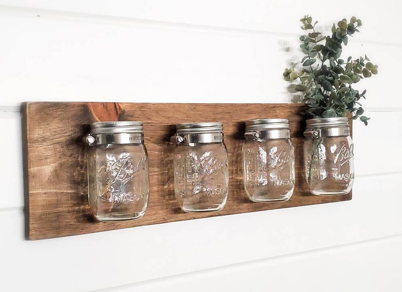 May include: A rustic wooden wall shelf with four clear glass mason jars attached with metal clips. The jars are empty and ready to be used for storage or decoration.