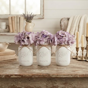 May include: Three white painted glass Ball Mason jars arranged in a row, each featuring the embossed Ball logo and filled with light purple hydrangea flowers. Each jar is tied with a thin brown twine bow around the neck of the glass container.