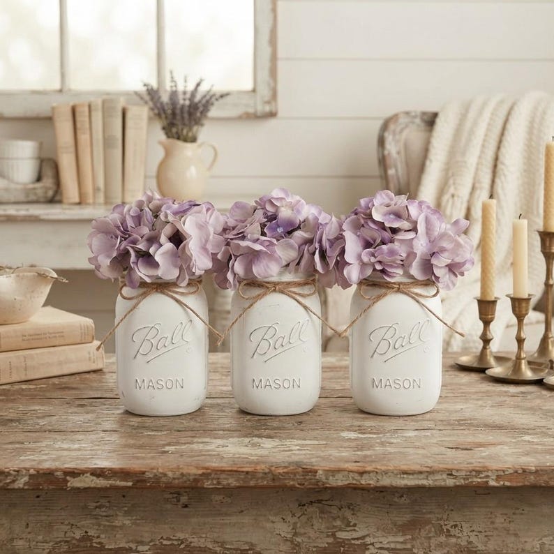 May include: Three white painted glass Ball Mason jars arranged in a row, each featuring the embossed Ball logo and filled with light purple hydrangea flowers. Each jar is tied with a thin brown twine bow around the neck of the glass container.