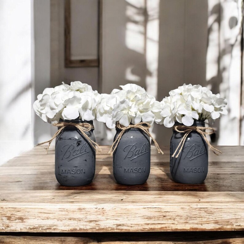 May include: Three matte dark gray glass Ball Mason jars, each filled with a cluster of white hydrangea flowers. Each jar features the embossed Ball Mason logo on the front and is tied around the neck with a thin, natural brown jute twine bow.
