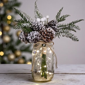 May include: A clear glass mason jar filled with fairy lights, pinecones, and faux snow-covered greenery. The jar is tied with twine and sits on a wooden surface, creating a festive holiday decoration.