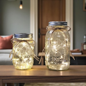 May include: Two clear glass Mason jars with silver lids, filled with fairy lights. Each jar is adorned with a brown twine bow. The jars are on a wooden surface, creating a warm, inviting ambiance. Home decor.