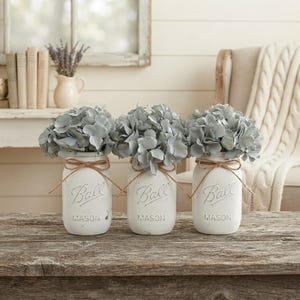 May include: Three white painted glass mason jars, each embossed with the word Ball and Mason, are filled with light blue hydrangea flowers. Each jar is tied with a thin brown twine bow around the neck, creating a set of decorative floral arrangements.