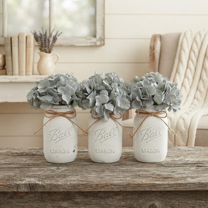 May include: Three white painted glass mason jars, each embossed with the word Ball and Mason, are filled with light blue hydrangea flowers. Each jar is tied with a thin brown twine bow around the neck, creating a set of decorative floral arrangements.