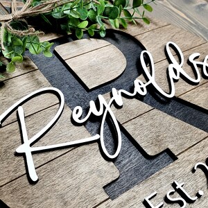 May include: A wooden sign with the last name "Reynolds" written in white cursive script. The letter "R" is cut out of the wood and painted black. The sign also includes the text "Est. 20..." in black.