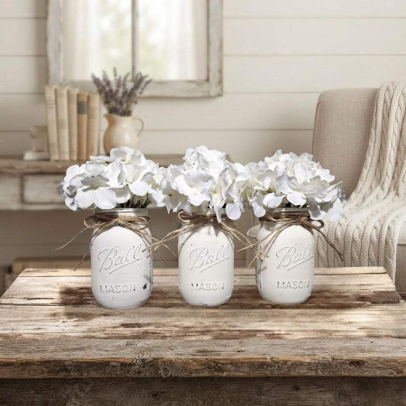May include: Three white painted glass Ball Mason jars, each filled with a bouquet of white hydrangea flowers. Each jar features the embossed Ball Mason logo and is tied with a thin brown twine bow around the neck, arranged in a row.