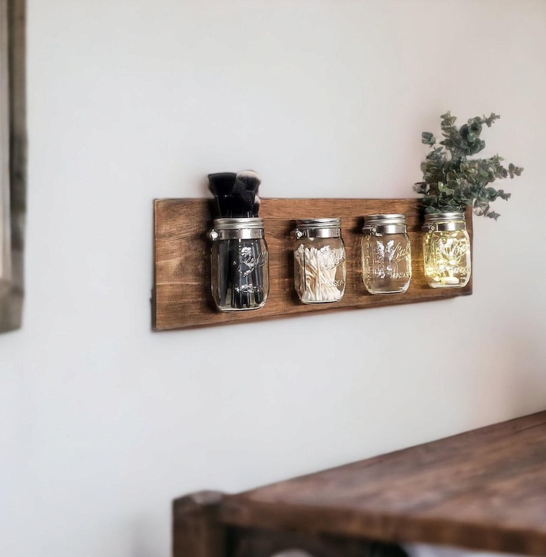 May include: A wooden shelf with four glass jars attached to it. The jars are filled with various items, including makeup brushes, cotton swabs, and decorative lights. The shelf is mounted on a white wall.
