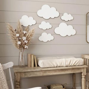 May include: A set of six white cloud-shaped wall decorations arranged on a horizontal paneled wall. The clouds vary in size and feature smooth, rounded edges, creating a cohesive display above a wooden table with a glass vase of dried plants.