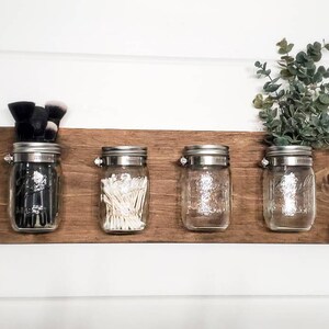 May include: A wooden wall organizer with four glass jars with metal lids. The jars are filled with various items, including makeup brushes, cotton swabs, and greenery.
