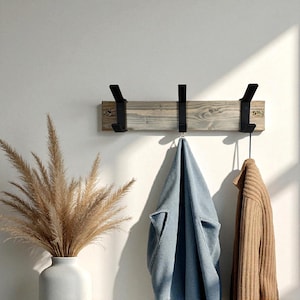 May include: A wooden wall-mounted coat rack with three black metal hooks. A light blue jacket and a tan sweater hang from the hooks. A white vase with dried ornamental grass is in the foreground.