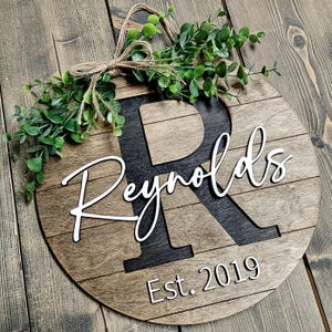May include: A round wooden sign with a black letter "R" and the text "Reynolds Est. 2019" in white. The sign is decorated with a green leafy garland.