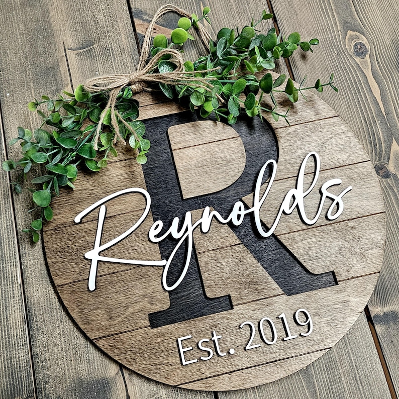 May include: A round wooden sign with a black letter "R" and the text "Reynolds Est. 2019" in white. The sign is decorated with a green leafy garland.