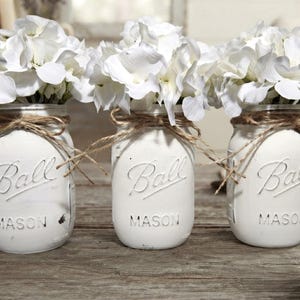 May include: Three white painted glass Ball Mason jars, each filled with a cluster of white hydrangea flowers. Each jar features the embossed Ball logo and the word MASON, with a piece of brown twine tied in a bow around the neck of the jar.