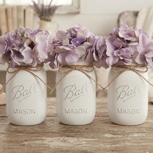 May include: Three white painted glass Ball Mason jars arranged in a row, each filled with light purple hydrangea flowers. Each jar features the embossed Ball logo and is decorated with a thin brown twine bow tied around the neck of the jar.