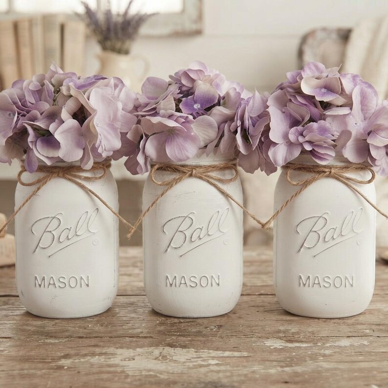 May include: Three white painted glass Ball Mason jars arranged in a row, each filled with light purple hydrangea flowers. Each jar features the embossed Ball logo and is decorated with a thin brown twine bow tied around the neck of the jar.