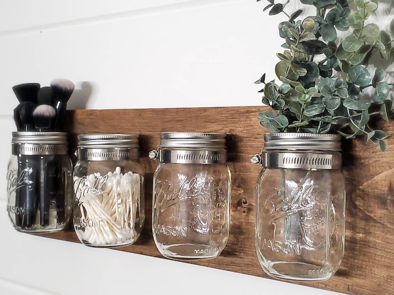 May include: Four clear glass mason jars with silver lids are mounted on a wooden shelf. The jars are filled with various items, including makeup brushes, cotton swabs, and greenery.