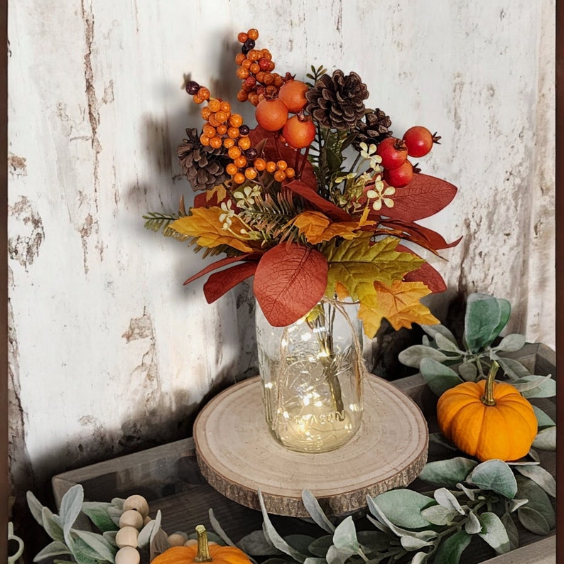 May include: Autumn-themed fall floral arrangement in a mason jar with string lights. The arrangement features artificial berries, pinecones, and leaves in shades of orange, red, and brown. Small pumpkins and greenery surround the base. The mason jar is labeled 'Mason'.