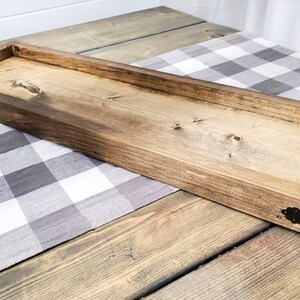 May include: A long, rectangular wooden tray with a natural wood finish. The tray has a raised edge and is decorated with metal corner accents. The tray is sitting on a grey and white checked tablecloth.