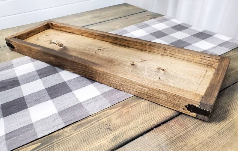 May include: A long, rectangular wooden tray with a natural wood finish. The tray has a raised edge and is decorated with metal corner accents. The tray is sitting on a grey and white checked tablecloth.