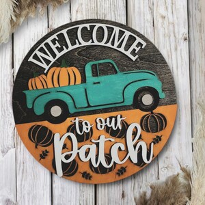 May include: Round wooden sign with a teal blue pickup truck carrying pumpkins. The sign says "Welcome to our Patch" with a fall-themed orange and brown background.