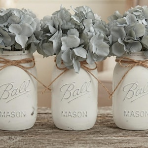 May include: Three white painted glass Ball Mason jars arranged in a row, each filled with light gray hydrangea flowers. Each jar features the embossed text Ball Mason and is tied with a thin brown twine bow around the neck of the glass container.