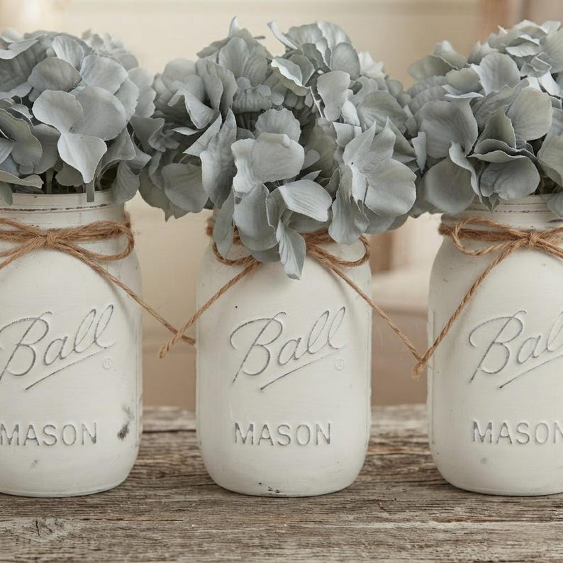 May include: Three white painted glass Ball Mason jars arranged in a row, each filled with light gray hydrangea flowers. Each jar features the embossed text Ball Mason and is tied with a thin brown twine bow around the neck of the glass container.